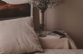 A calm, dimly lit bedside scene at night: a bed with a cream pillow, a vase of dried flowers, and a phone charging on the nightstand.