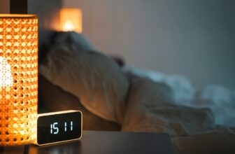 A digital alarm clock on a nightstand beside a bed in warm, dim evening light