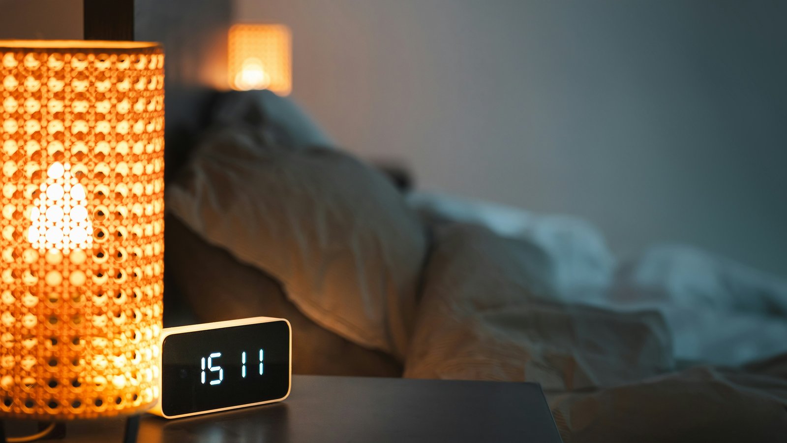 A digital alarm clock on a nightstand beside a bed in warm, dim evening light