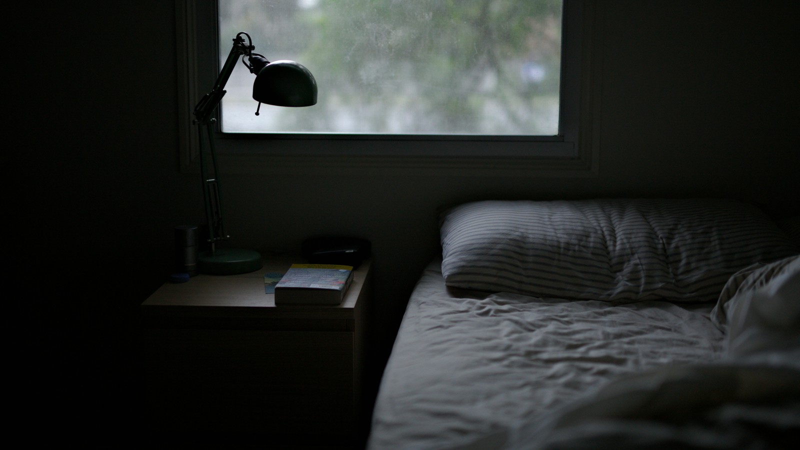 A dark, cool bedroom at dusk with a bed, nightstand lamp, and a window — set up for better sleep