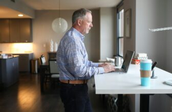 A man working while standing at a standing desk in a bright home, with a laptop and coffee — moving more during the workday.