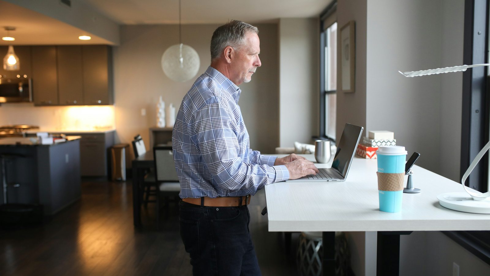 A man working while standing at a standing desk in a bright home, with a laptop and coffee — moving more during the workday.