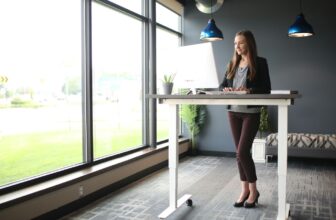 A woman working while standing at a height-adjustable standing desk beside large office windows with greenery outside.