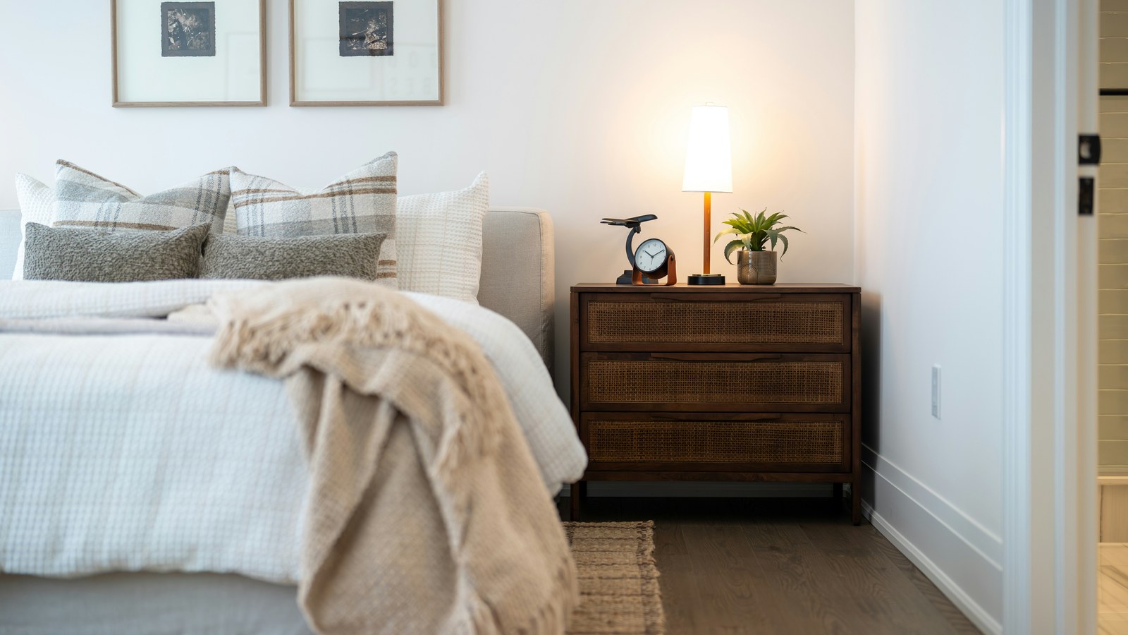 A tidy bedroom with a made bed, a lit bedside lamp, an analog alarm clock, and a small potted plant on a wooden nightstand.