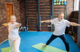 An older man and woman doing a gentle standing yoga stretch in a bright studio