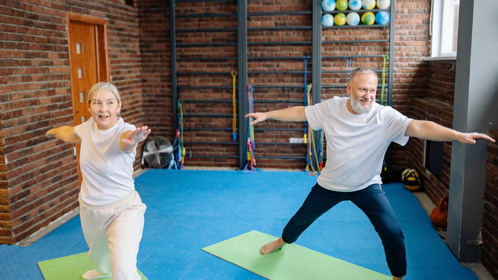 An older man and woman doing a gentle standing yoga stretch in a bright studio