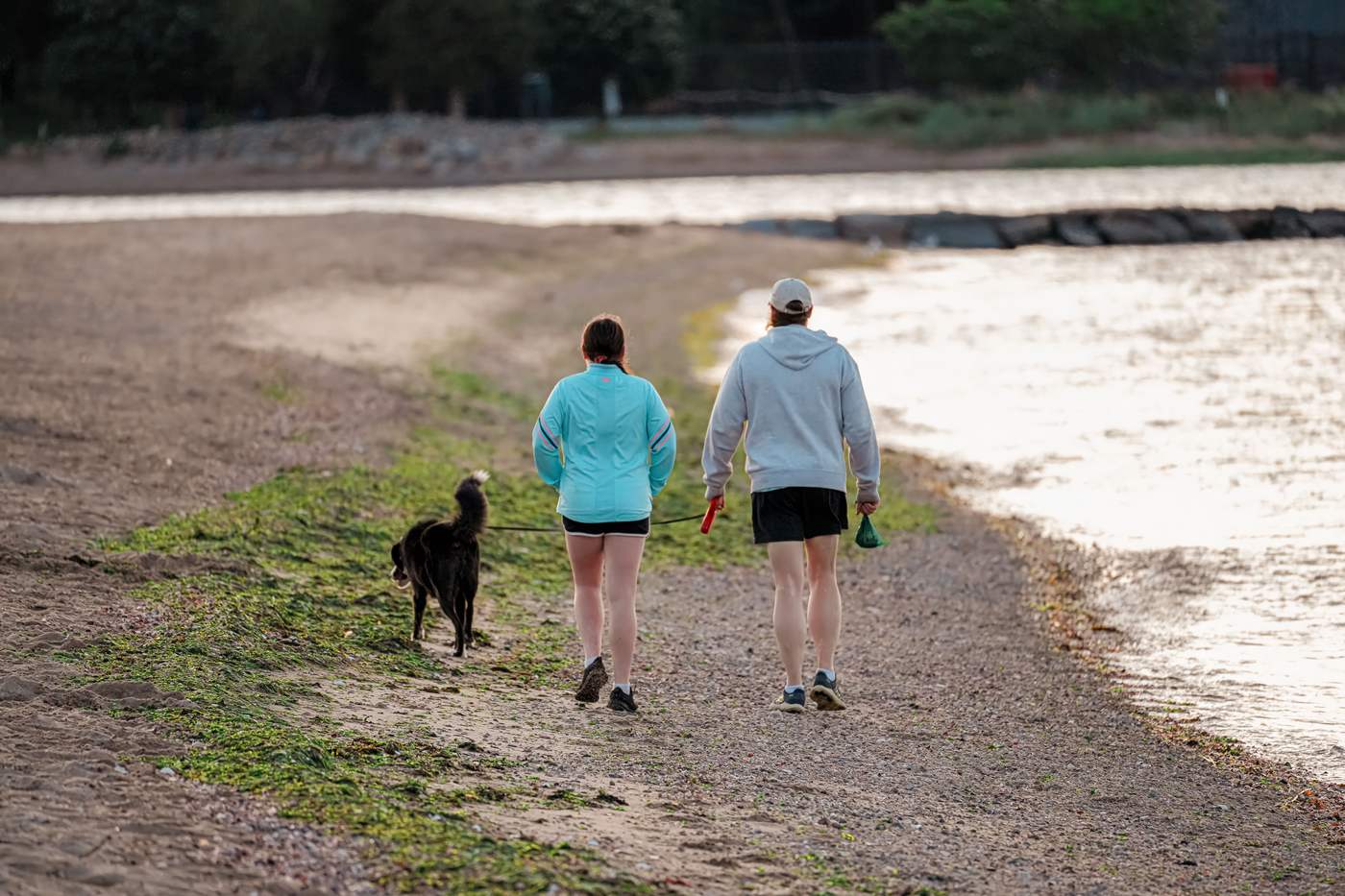 Two people walking a dog along a gravel path beside water